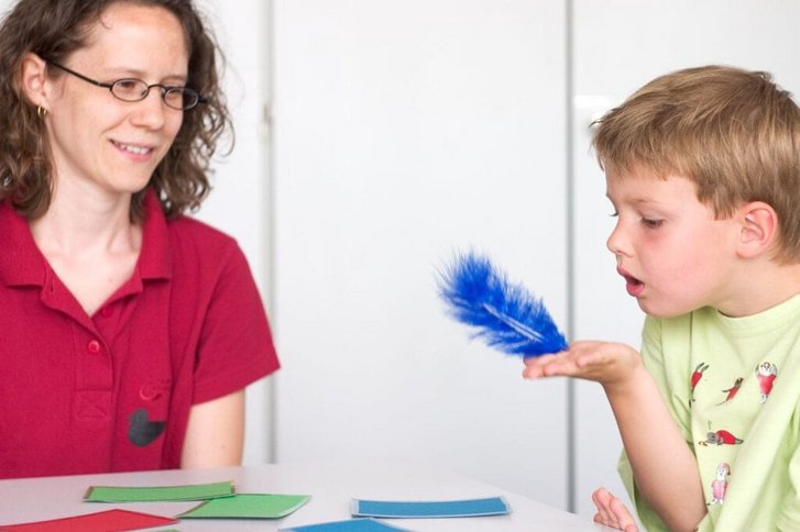 Kind pustet auf eine auf seiner Hand liegenden blauen Feder unter Anleitung der Logopaedin