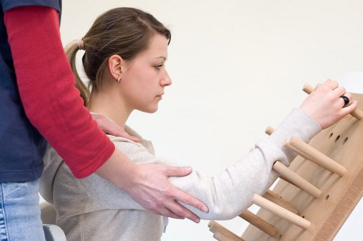 Eine Patientin uebt mit Unterstuetzung einer hinter ihr stehenden Therapeutin Greifen an einem schraegen Brett mit Holzstaeben in der Handtherapie im VAMED Rehazentrum Karlsruhe