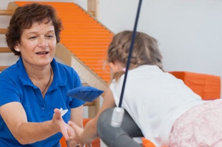 Ein Kind liegt baeuchlings auf einer Schaukel und uebergibt der frontal abgebildeten Therapeutin einen Ball in der Kinderbehandlung der Ergotherapieabteilung des VAMED Rehazentrums Karlsruhe