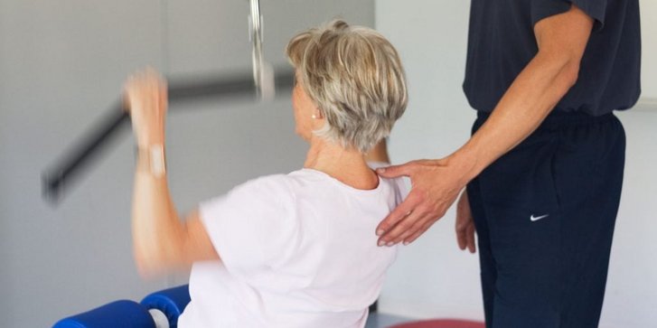 Patientin im KG am Gerät die bei der Behandlung in der Physiotherapie vom Physiotherapeuten unterstützt wird