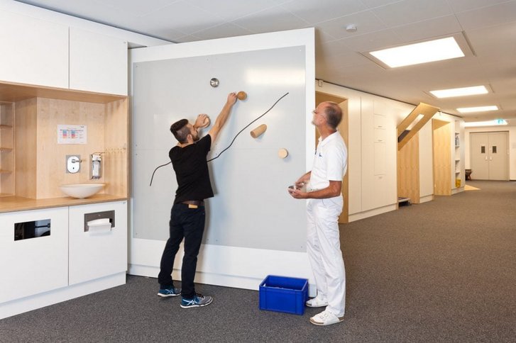 Ein Patient steht an einer Wand und uebt mit einem Therapeuten weite Greifbewegungen bei der arbeitsplatzspezifischen Trainingstherapie in der Reha