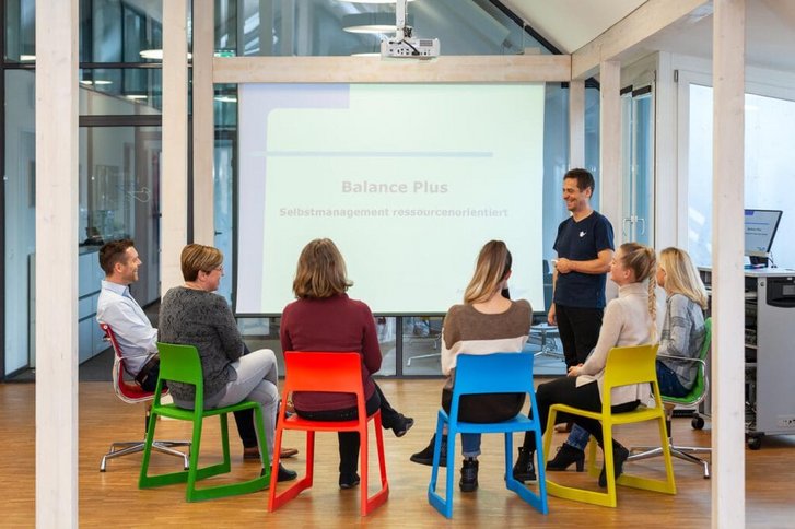 Sicht von hinten auf sechs lachende im Halbkreis vor einer Projektionsleinwand sitzende Menschen die einen Vortrag zu Stressbewältigung bekommen