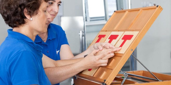 Ergotherapeutin fuehrt beim Wahrnehmungstraining mit einer Patientin deren Finger ueber rote hervorgehobene Buchstaben