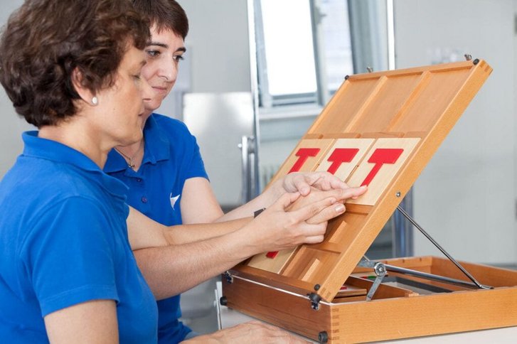 Ergotherapeutin fuehrt beim Wahrnehmungstraining mit einer Patientin deren Finger ueber rote hervorgehobene Buchstaben