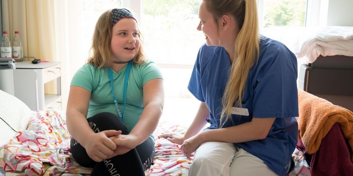 Maedchen sitzt auf Bett und spricht mit Pflegekraft