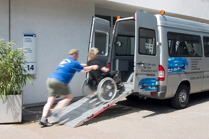 Fahrdienstmitarbeiter schiebt Patient im Rollstuhl über eine Laderampe ins Auto