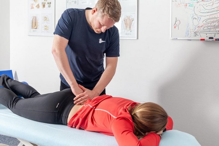 Ein Physiotherapeut arbeitet über Weichteiltechniken am Lendenbereich einer in Bauchlage liegenden Patientin