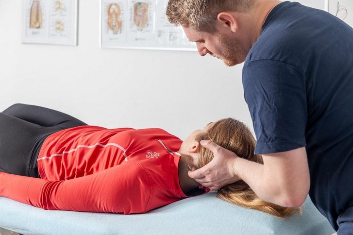 Ein Therapeut untersucht den Kopf einer Patientin in der physiotherapeutischen Behandlung die in Rueckenlage auf einer Liege liegt