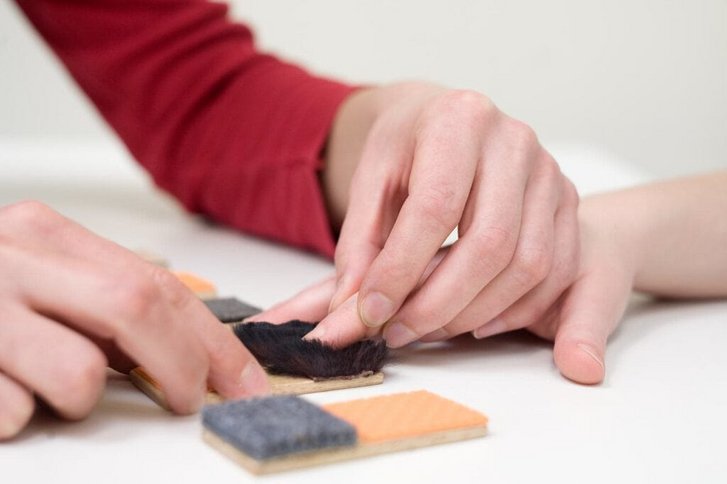 Nahaufnahme von Haenden einer Ergotherapeutin die im Rahmen der Handtherapie den Finger einer Patientin ueber verschiedene Oberflaechen fuehrt